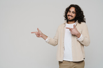 Smiling hispanic man pointing finger away at copy space while standing isolated over white wall