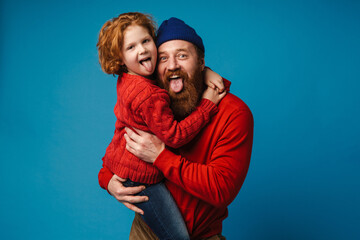 Cheerful father and son hugging and sticking their tongues out isolated over blue background