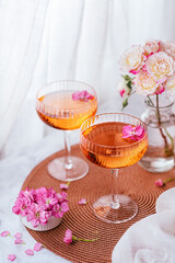 Two cocktails decorated with flowers on a white table next to the window and a vase with roses in the background.