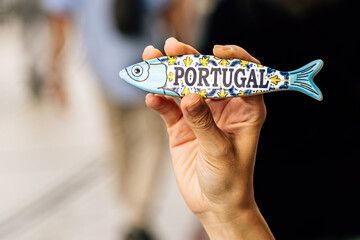 Hand of a young woman holding a sardine-shaped magnet on the street. Souvenir concept from Lisbon,...