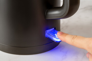 Woman finger turning on the switch to boil kettle. Switch glows blue while powered on. Matte black...