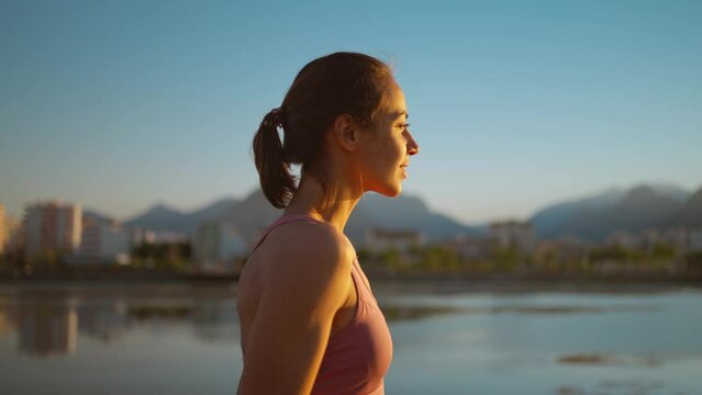 portrait calm confident woman standing on wooden jetty pier, enjoying watching sunset with mountains and sea view. woman relaxing after evening workout, breathing exercises, yoga and meditation