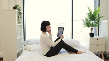 Side view of caucasian woman holding phone while looking at online website with trendy clothes on sale using digital tablet. Casually dressed lady enjoying shopping with big discount at home.