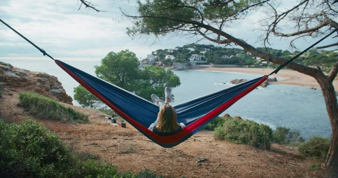 Woman sways in a hammock on a green hill, surrounded by trees and forest. Her gaze rests on the serene blue sea, while she relaxes and meditates in the summer breeze.