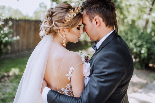 A stylish young groom in a suit and a beautiful blonde model bride with a diadem, a crown on her head are gently hugging outdoors. Close-up wedding photography, portrait of the newlyweds.