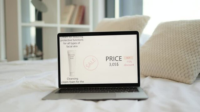 Portable laptop placed on white bed with cosmetic ecommerce site on screen. Price for cleansing cream foam with discount of 15 percent is 3,05 dollars. Seasonal shopping during sale.