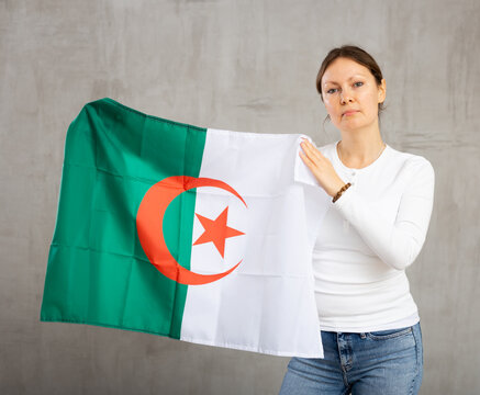 Calm female with serious expression on face standing against gray wall with national flag of Algeria