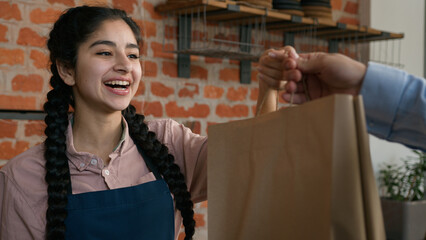 Friendly young indian woman waitress barista giving takeaway food bag to customer female seller cafe employee in apron holding takeout order delivery in cafeteria restaurant coffee shop serves client