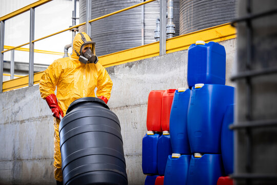 Experienced worker in protection suit and gas mask moving barrels with sulfuric acid inside chemicals production plant.
