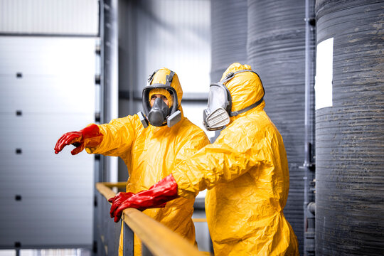 Factory worker standing by large metal storage tanks with acids wearing yellow protection suit, gas mask and gloves explaining trainee process of chemicals production inside the plant.