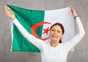 Portrait of happy woman posing with national flag of Algeria in hands on gray background in studio