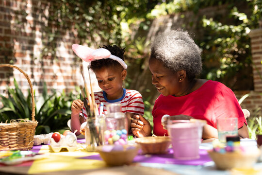 Happy african american grandmother and grandson coloring easter eggs in garden - Powered by Adobe