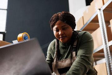 Cheerful supervisor analyzing merchandise logistics on laptop computer while preparing customers...