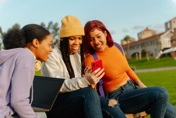 multiracial group of 3 female friends sitting on a bench on a university campus using a cell phone...
