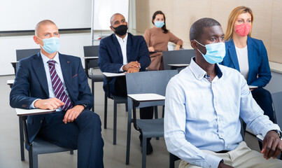 International group of business people wearing protective face masks listening to presentation in conference room. Concept of precautions and social distancing in COVID 19 pandemic..