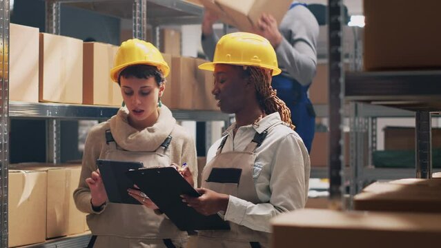 Multiethnic team of supervisors counting boxes of goods placed on warehouse racks, looking at merchandise products in packages. Young women in overalls working in storage room.