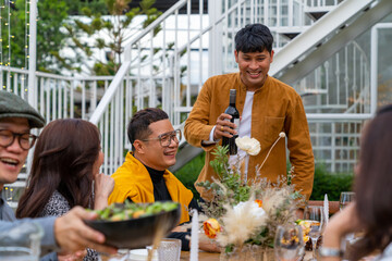 Asian man pouring red wine into wine glass to friends during outdoor celebration dinner party in the garden on summer holiday vacation. Man and woman friends reunion dining meeting party at restaurant