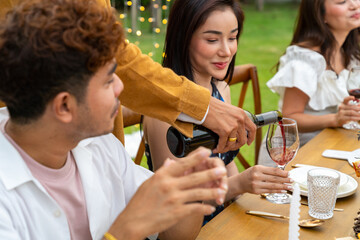 Asian man pouring red wine into wine glass to friends during outdoor celebration dinner party in the garden on summer holiday vacation. Man and woman friends reunion dining meeting party at restaurant