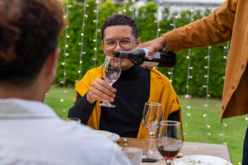Asian man pouring red wine into wine glass to friends during outdoor celebration dinner party in the garden on summer holiday vacation. Man and woman friends reunion dining meeting party at restaurant
