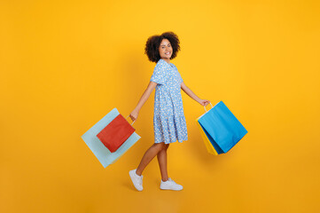 Full length photo of a lovely cheerful curly haired african american woman dressed in a blue summer dress, holding shopping bags, looking at camera, smiling, stand on isolated pastel pink background