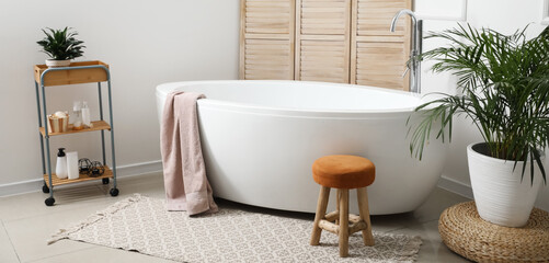 Modern bathtub with cosmetics and houseplant in light interior