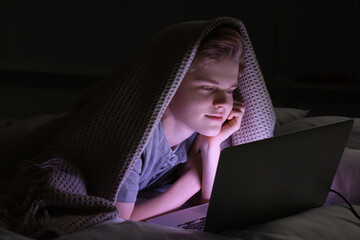 Teenage boy using laptop under blanket on bed at night. Internet addiction