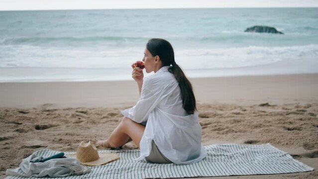 Woman relaxing beach picnic sitting at blanket. Girl enjoying fruits on seashore