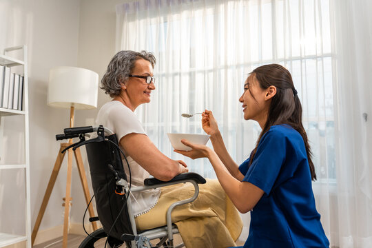 Asian Caregiver Nurse Serve Food To Caucasian Senior Older Woman At Home. 