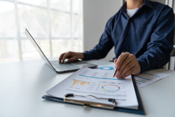 young business man Recording data on laptop, comparing details from graphs. Business income charts in the areas of real estate, tax, management, technology marketing in a private office.