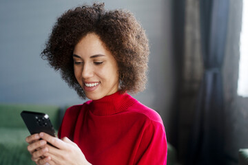 Smiling curly haired African American woman holding mobile phone reading text message, communication online at home. Stylish female shopping online, copy space. Technology concept  