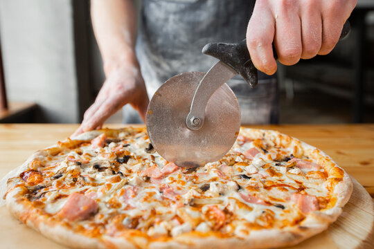 Closeup hand of chef baker in uniform blue apron cutting pizza at kitchen
