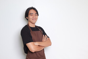 Portrait of attractive Asian barista man in brown apron keeping arms crossed and looking at camera. Advertising concept. Isolated image on white background - obrazy, fototapety, plakaty