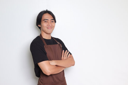 Portrait of attractive Asian barista man in brown apron keeping arms crossed and looking at camera. Advertising concept. Isolated image on white background