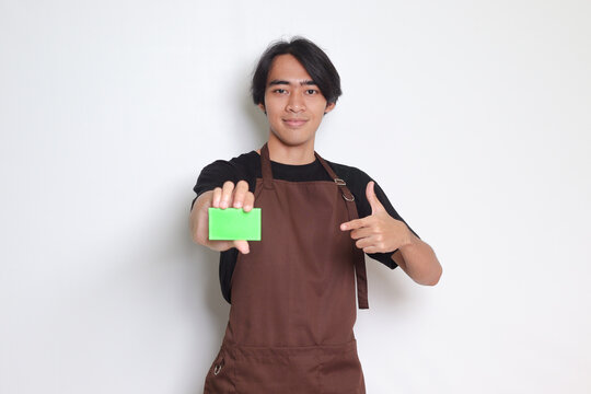 Portrait of attractive Asian barista man in brown apron showing template of credit card and pointing on it. Isolated image on white background