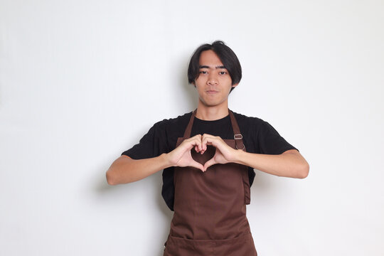 Portrait of attractive Asian barista man in brown apron speaks about own feelings, makes heart gesture over chest, expresses sympathy and love, smiles positively. Isolated image on white background