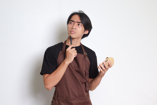 Portrait of attractive Asian barista man in brown apron taking order, writing on menu book list, thinking with pen on chin. Isolated image on white background