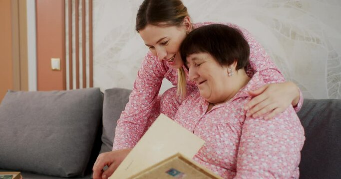Senior mom and adult daughter are singing a song looking at family photo album. They are sitting on sofa in living room at home wearing pink pajamas family look style. Memories, relationships, values.