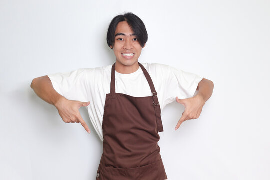 Portrait of attractive Asian barista man in brown apron showing product, pointing at something with hands. Advertising concept. Isolated image on white background