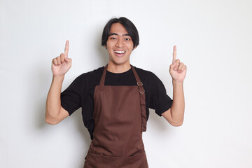 Portrait of attractive Asian barista man in brown apron showing product, pointing at something with...
