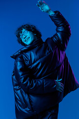 Obraz premium Man in winter jacket model poses and dances, hipster lifestyle, portrait blue background, mixed neon light, fashion style and trends teenager, copy space