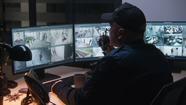 Security officer controls CCTV cameras in office, uses computers, talks by walkie-talkie. High tech software with surveillance cameras footage on screens. Modern security system and social safety.