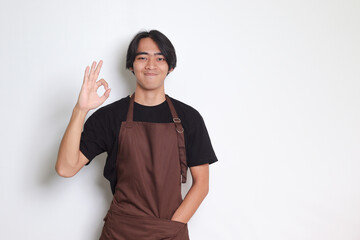 Portrait of attractive Asian barista man in brown apron showing ok hand gesture and smiling looking at camera. Advertising concept. Isolated image on white background