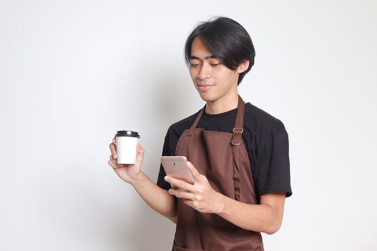 Portrait of attractive Asian barista man in brown apron holding disposable paper coffee cup while using mobile phone. Isolated image on white background