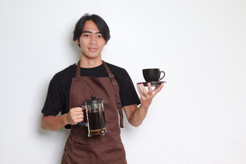 Portrait of attractive Asian barista man in brown apron showing a cup and saucer while holding...