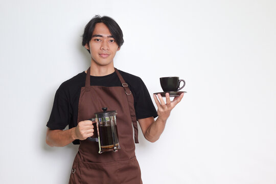 Portrait of attractive Asian barista man in brown apron showing a cup and saucer while holding french press coffee maker. Isolated image on white background