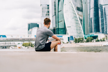Tired Young man runner sitting on stairs and relaxing after sport training. Holding water bottle while doing workout in summer city street