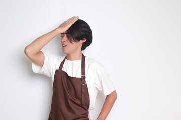 Portrait of attractive Asian barista man in brown apron holding hand on forehead, forgets something, and regrets himself. Isolated image on white background