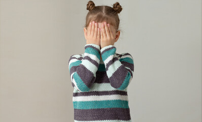Child placing hands across face to express embarrassment dismay or fear on grey background, caucasian little girl of 6 7 years in green gray striped sweater - obrazy, fototapety, plakaty