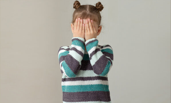 Child placing hands across face to express embarrassment dismay or fear on grey background, caucasian little girl of 6 7 years in green gray striped sweater