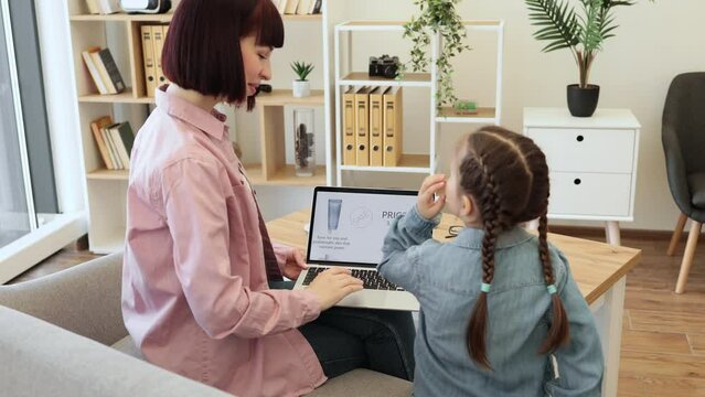 Rear view of beautiful woman holding laptop and cute girl looking over shoulder at her mom. Young females discussing facial beauty products provided at big discounts in online cosmetics webstore.